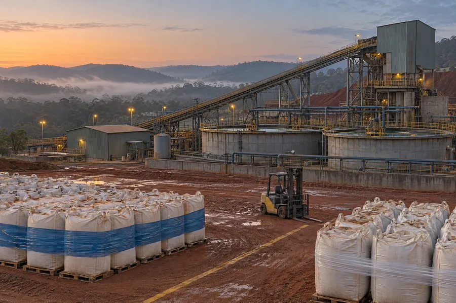 Pátio de planta de beneficiamento de minério ao amanhecer, com big bags de concentrado paletizados, empilhadeira, tanques e correia transportadora: o drawback intermediário na cadeia da mineração