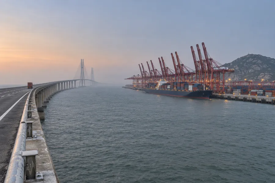 Buque cargado de contenedores atracado bajo las grúas de un puerto chino, con un puente sobre el mar al amanecer: la exportación de China a Brasil
